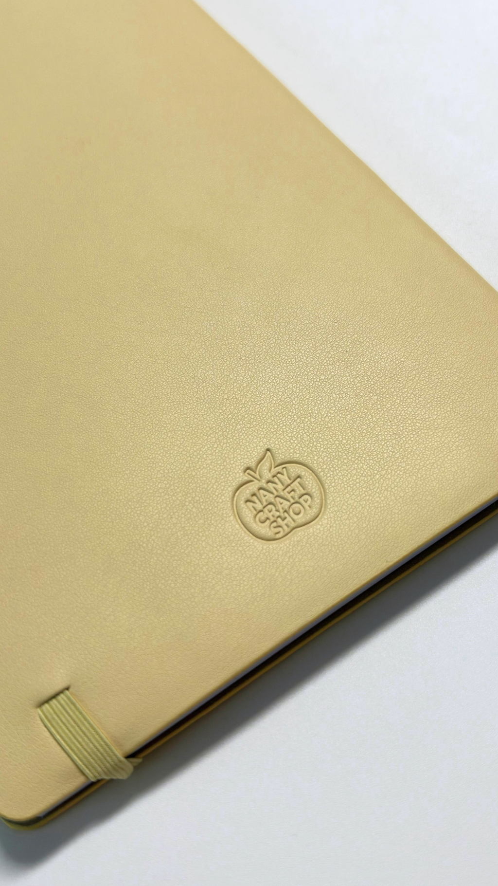 Gros plan sur un carnet beige doré à couverture texturée avec logo embossé et élastique de fermeture signé Nany Craft Shop dans la collection tous les produits