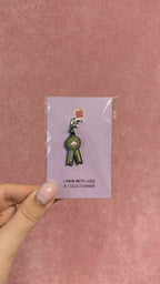 Charm métallique en forme de rosette noire et dorée présenté sur une carte rose tenue à la main par Nany Craft Shop